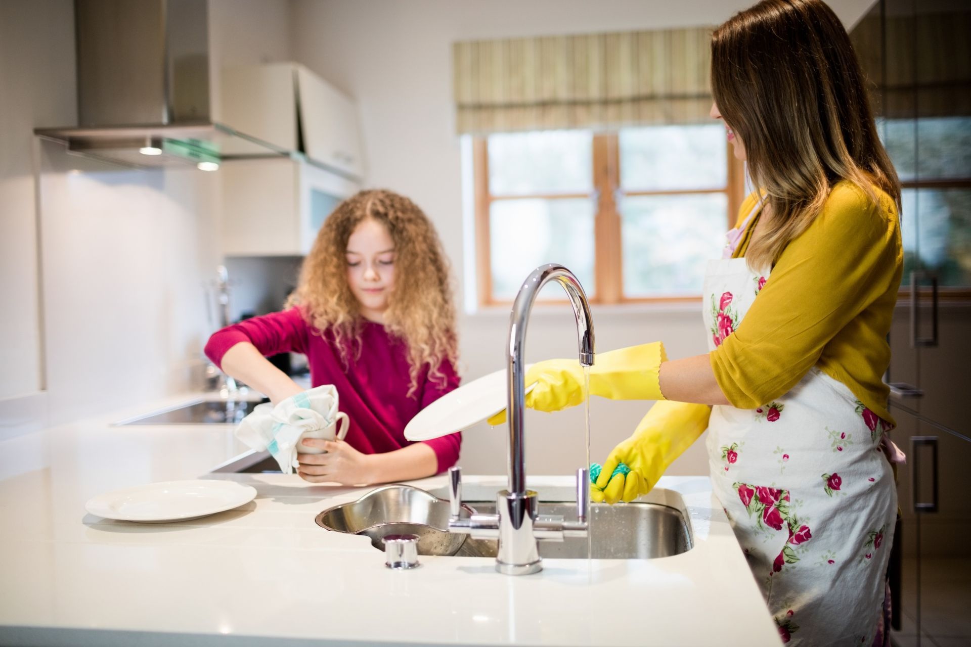 Woman and girl washing dishes in a kitchen. Girl dries a cup, woman scrubs with gloves.