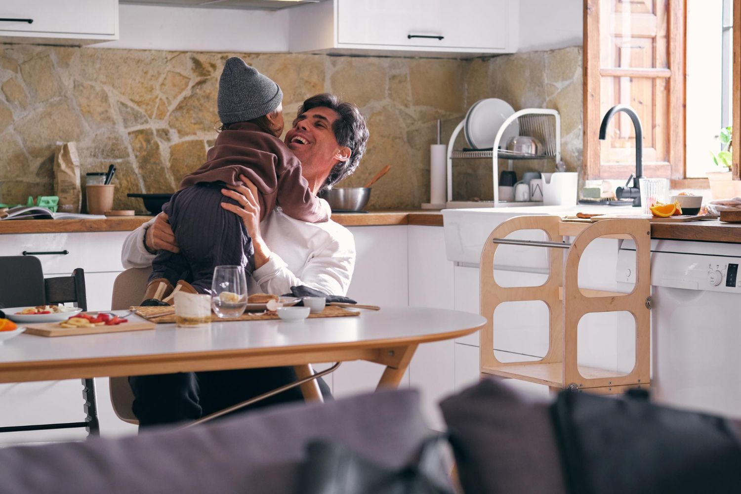 Man holding up child in a kitchen, smiling. Table with food; learning tower nearby.