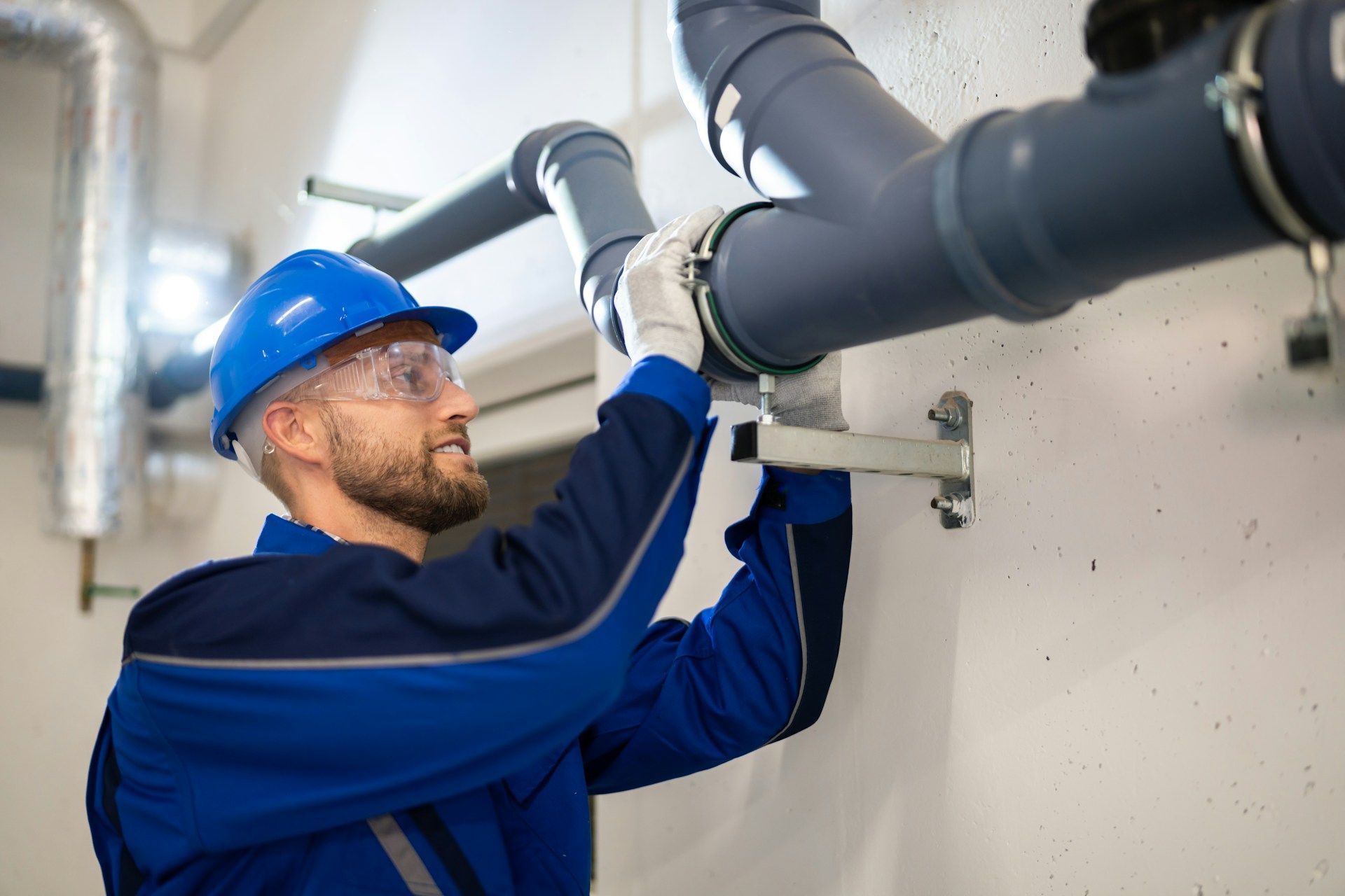 Plumber in blue uniform and hard hat, working on gray pipes mounted to a white wall.