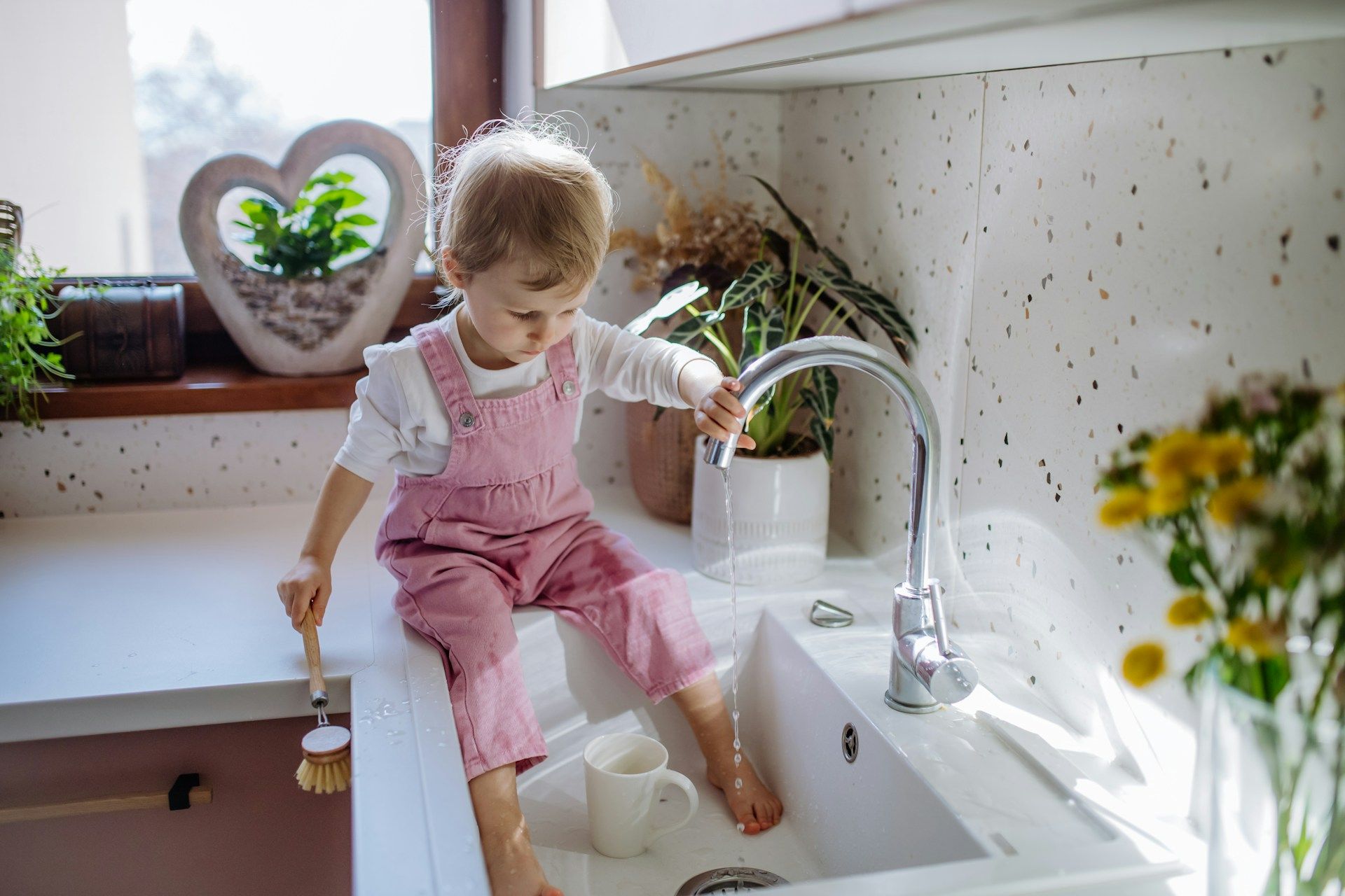 Child sits in kitchen sink, playing with water and a brush. Wearing pink overalls.