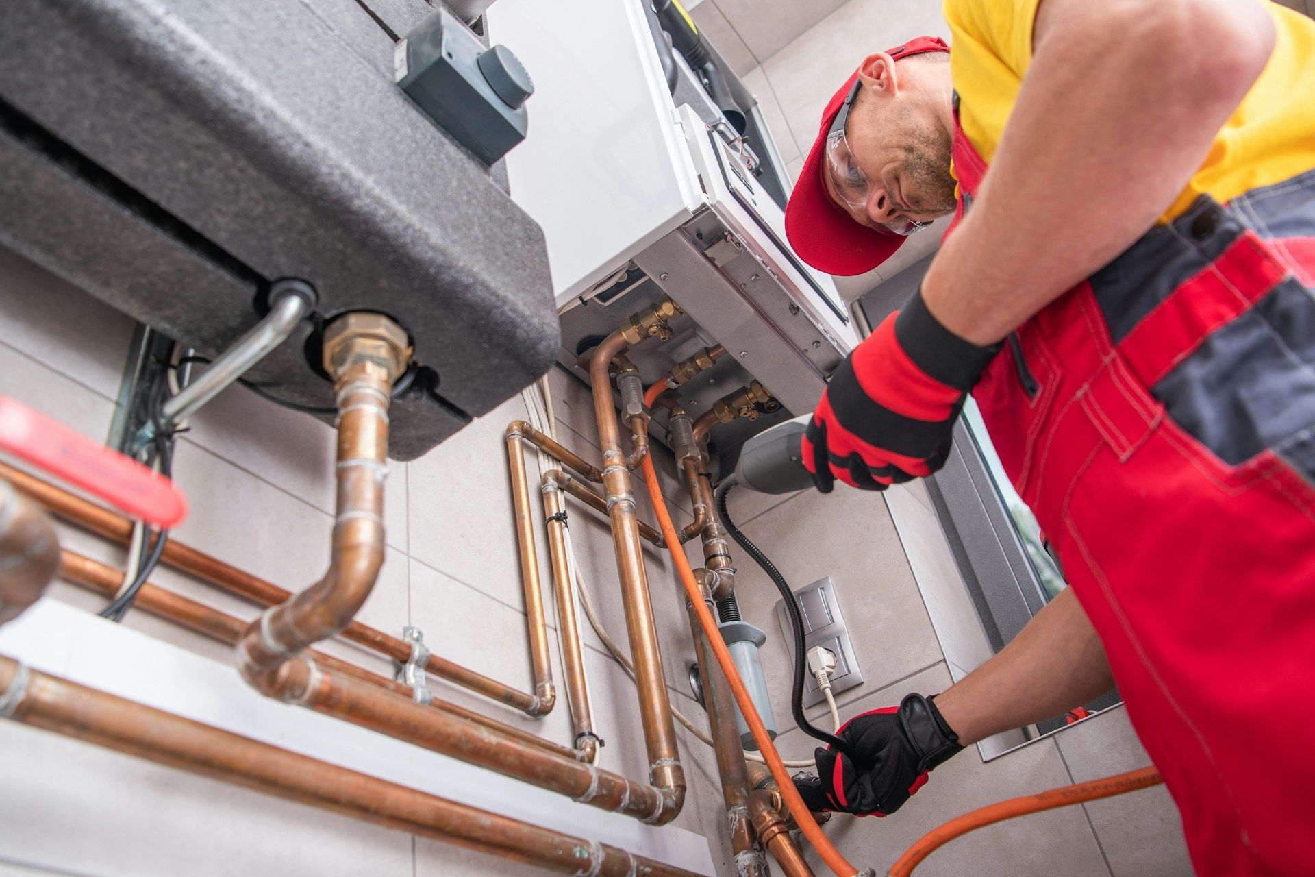 Plumber in red overalls inspecting copper pipes on a wall-mounted heating system, indoors.