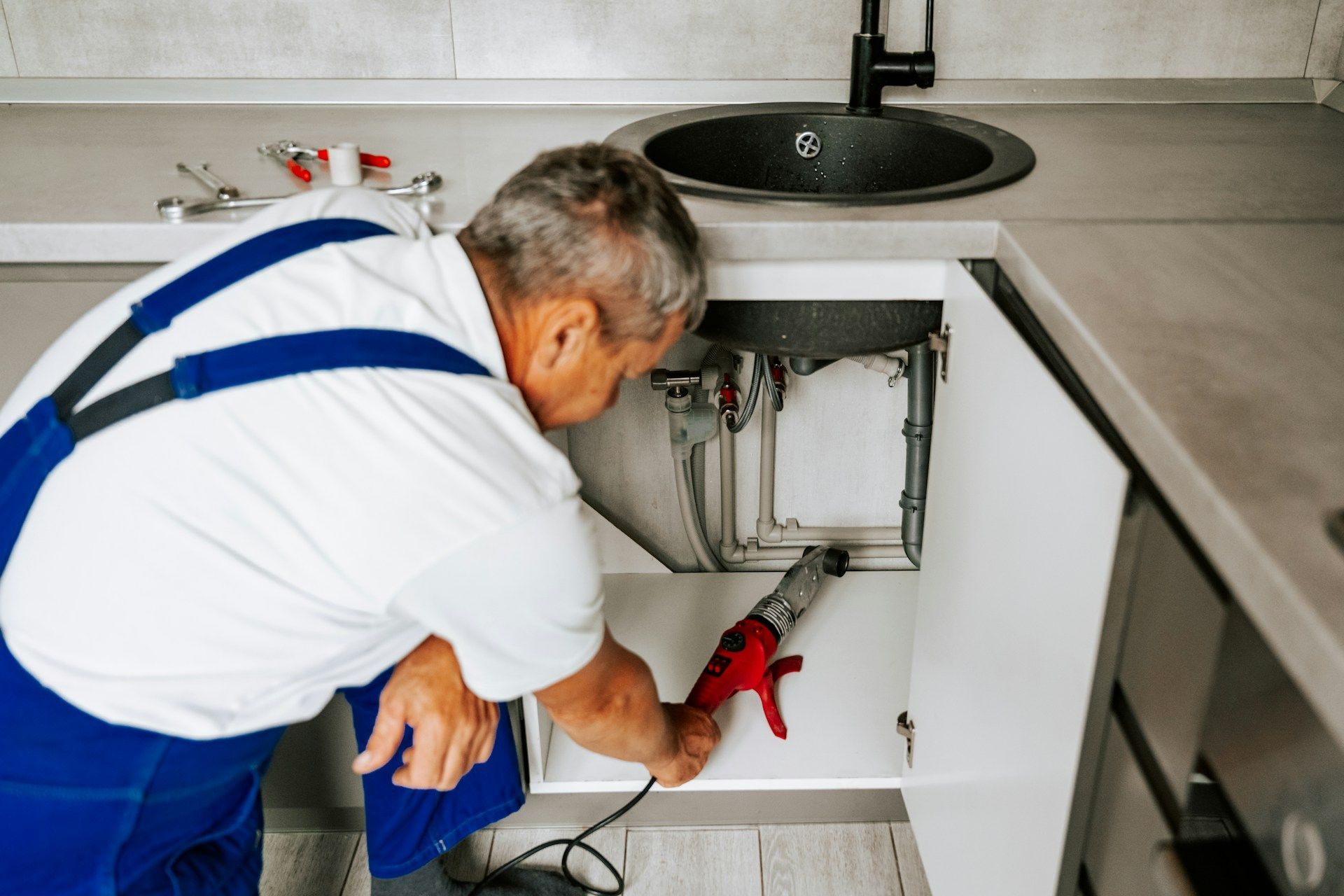 Plumber working under a kitchen sink, using a tool. Wearing blue overalls and white shirt.