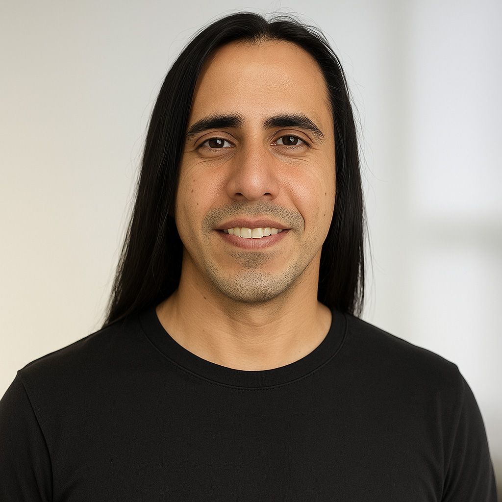 Man with long dark hair smiles at the camera, wearing a black shirt, against a blurred white background.