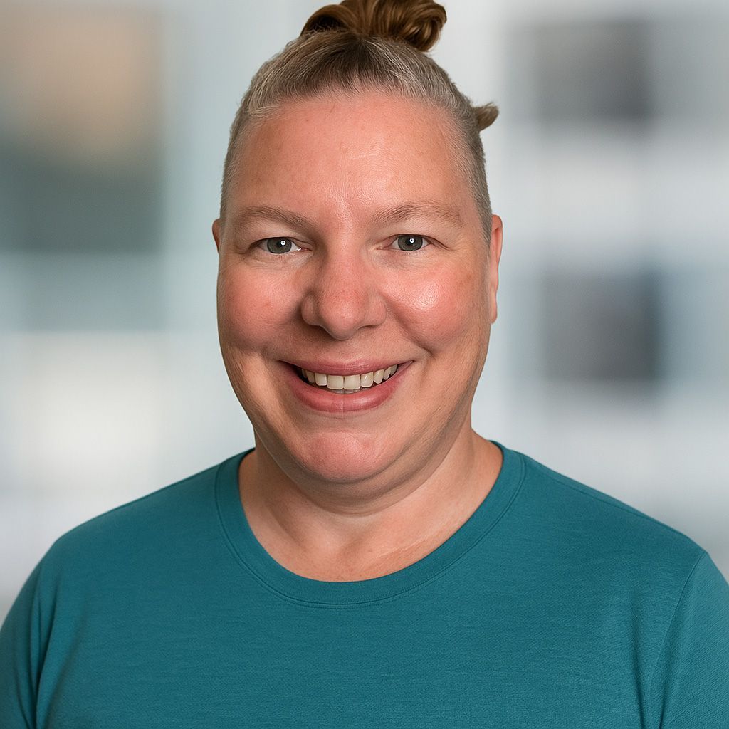 Smiling person with a topknot wearing a teal shirt. Background is a blurred office space.
