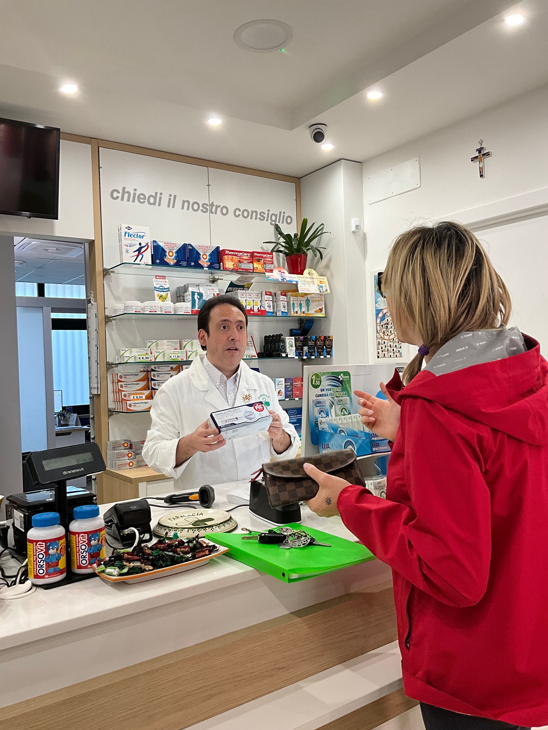 Un uomo in camice parla con una donna in giacca rossa al bancone di una farmacia.