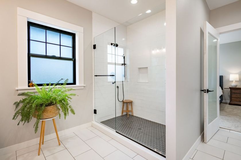 Modern bathroom with glass shower, white tile, black-framed window, and fern plant. Doorway to bedroom.