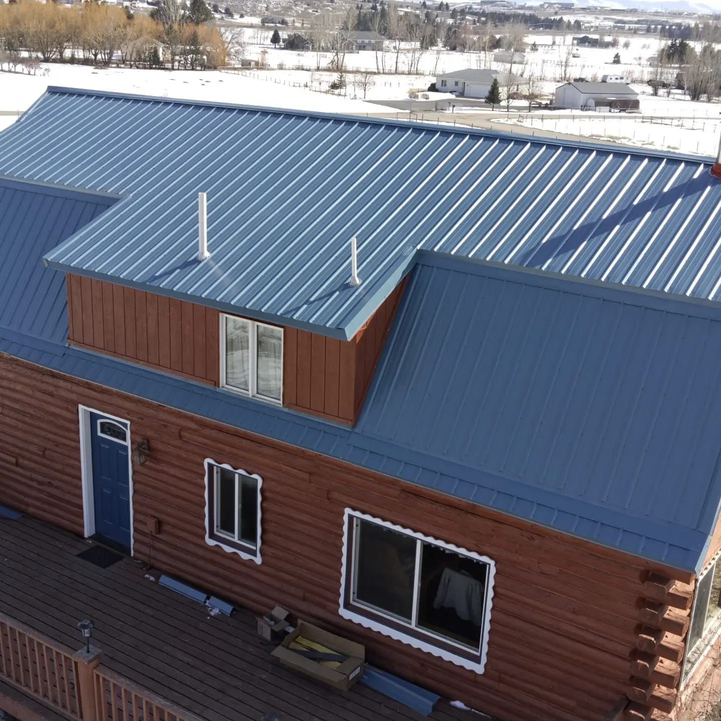 Log cabin with blue metal roof and snow-covered landscape in the background.