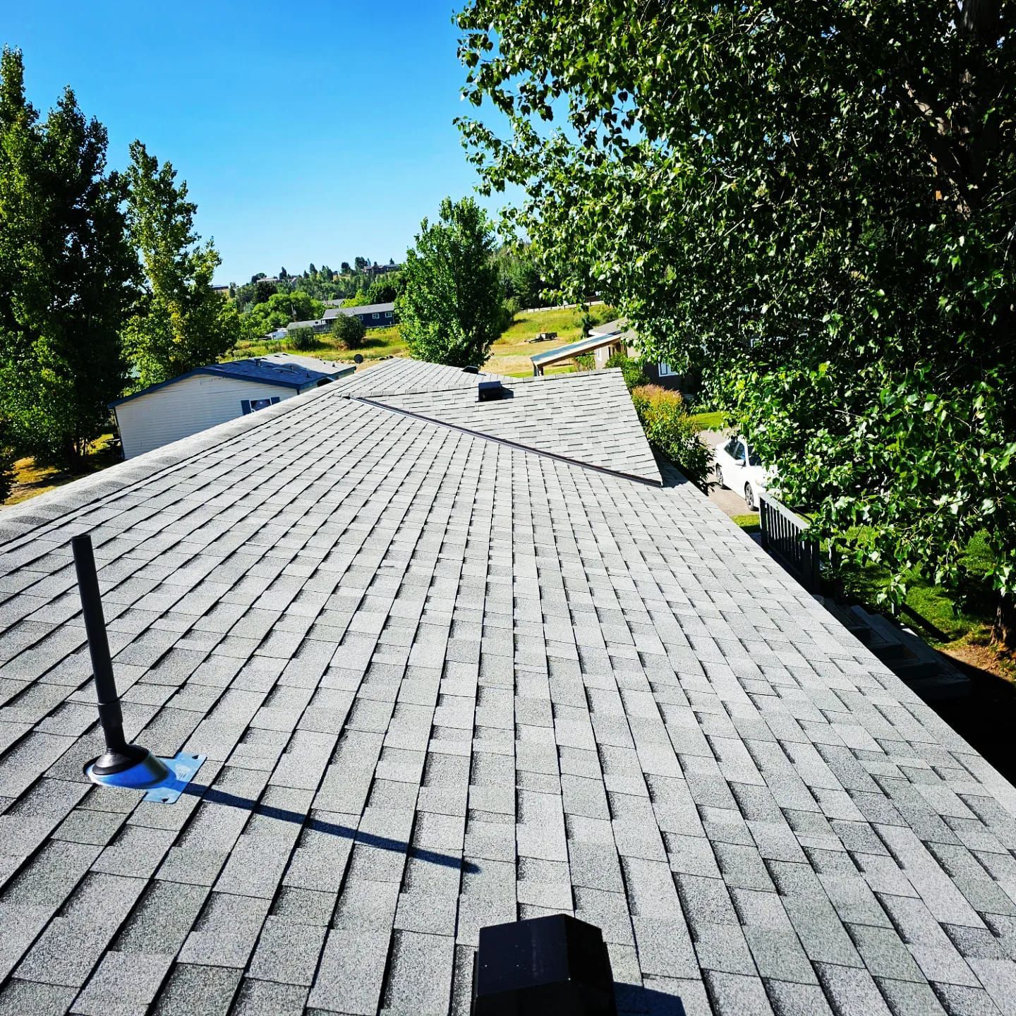Gray shingled roof with a vent pipe on a sunny day with trees in the background.