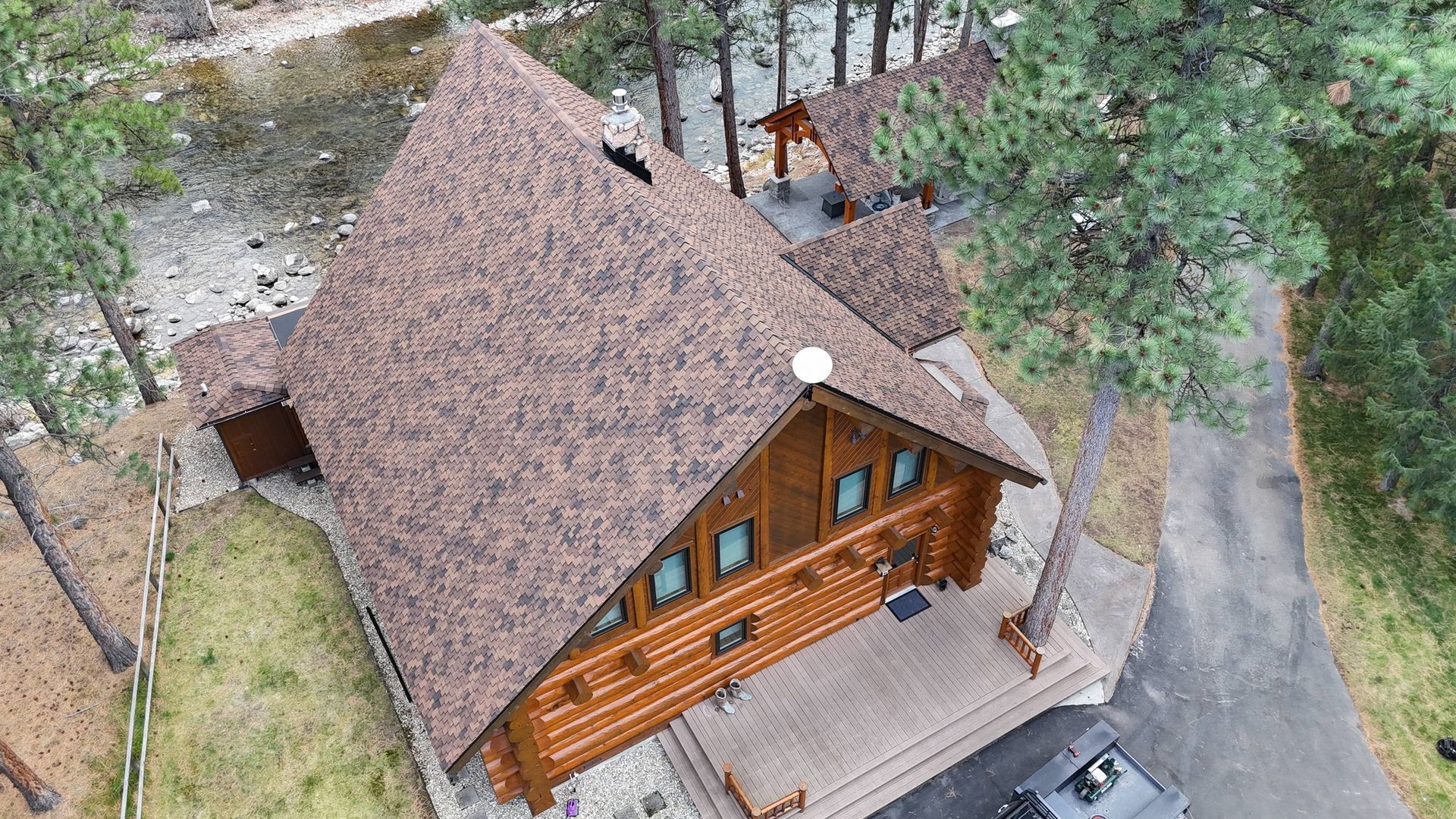 Aerial view of a log cabin with a brown roof surrounded by trees and a driveway.