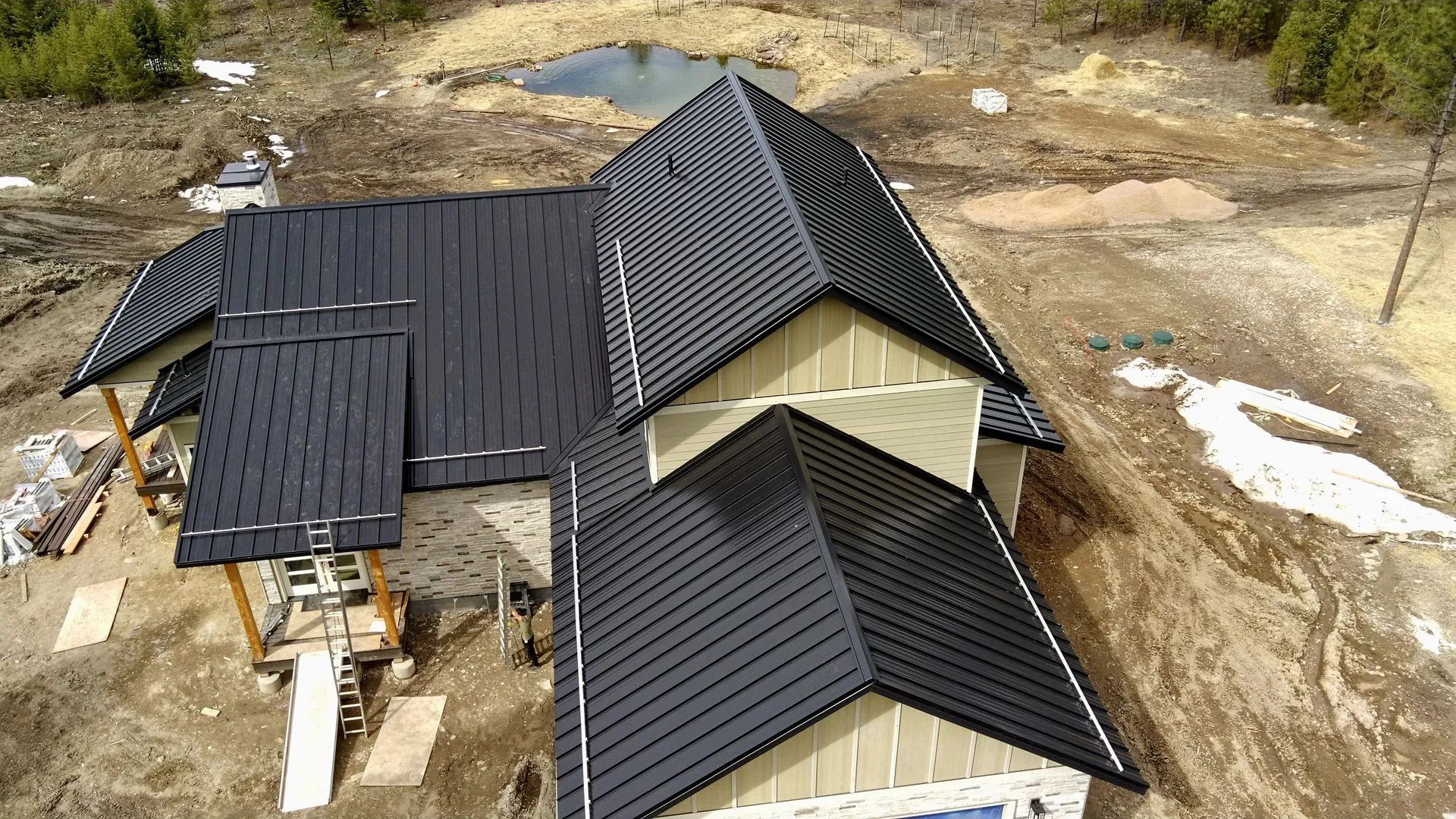 An aerial view of a house under construction with a newly installed black metal roof surrounded by dirt.