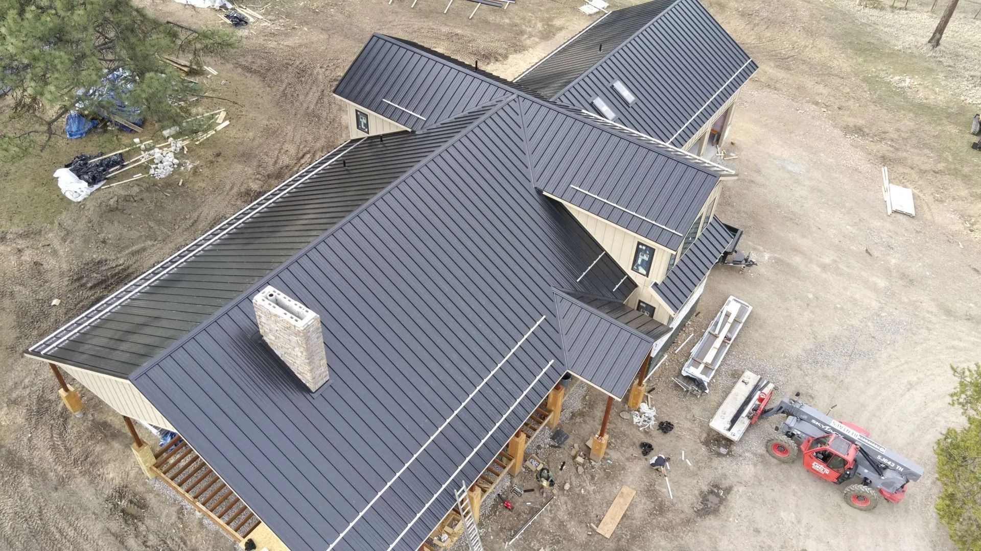 Aerial view of a house with a dark metal roof, construction equipment, and a dirt setting.