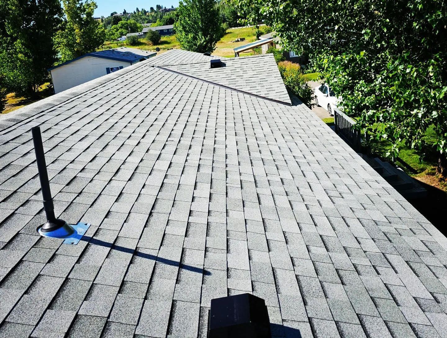 Gray asphalt shingle roof on a house, with a black vent pipe and chimney, trees in the background.