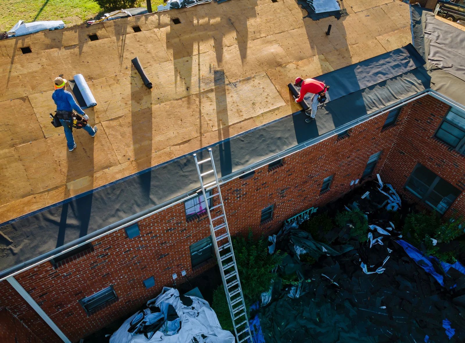 Two roofers working on a brick building's flat roof. One unrolls material; the other applies it. A ladder stands ready.