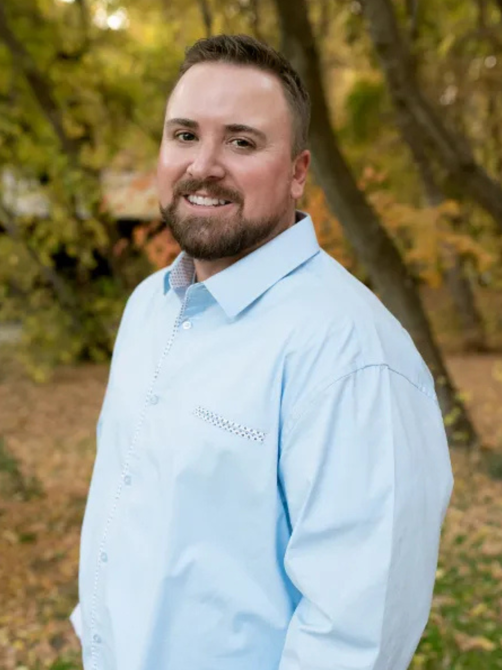 A man wearing a hat and a white shirt is smiling in front of a pleasant grove lindon chamber of commerce banner