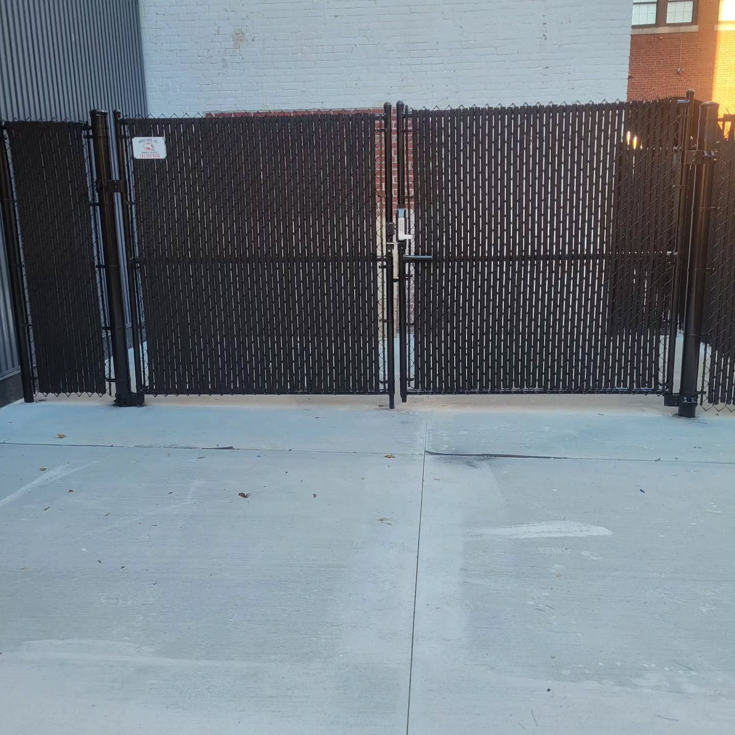 Black chain-link fence with gate, on a concrete surface. A light brick building is behind it.