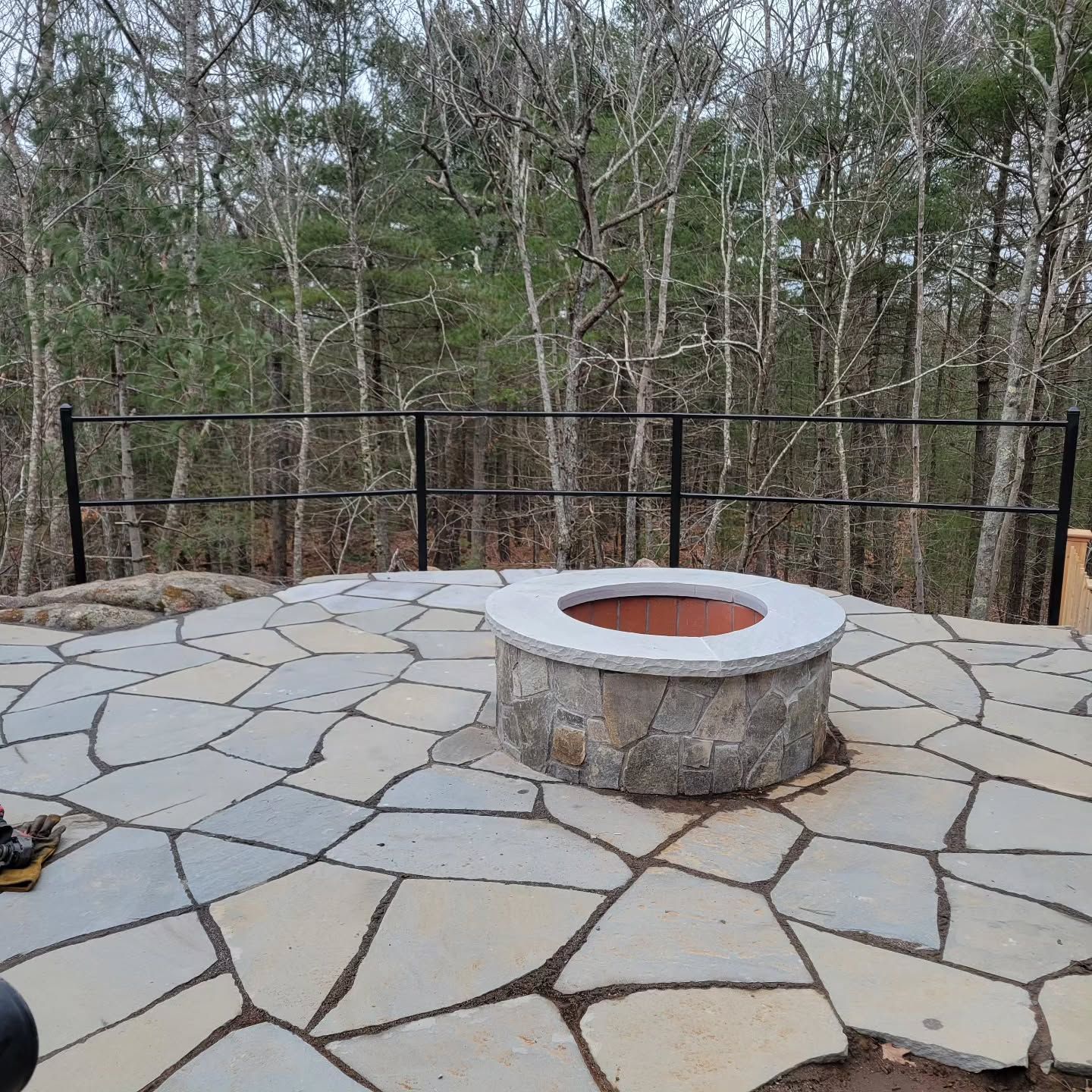 Stone patio with fire pit and black railing overlooking a forest.