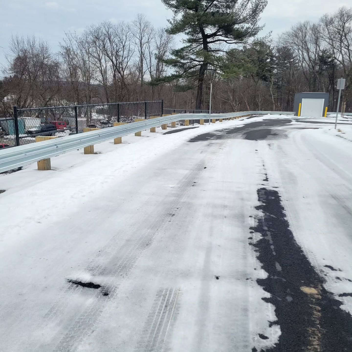 Snow-covered road with metal guardrail. Trees in the background, a small building to the right.