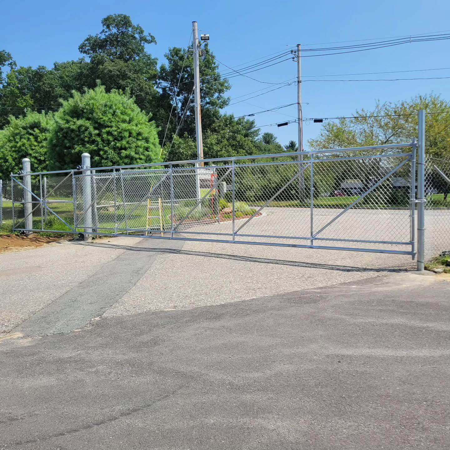 Chain-link fence gate, closed, on gravel drive. Trees and power lines in the background on a sunny day.