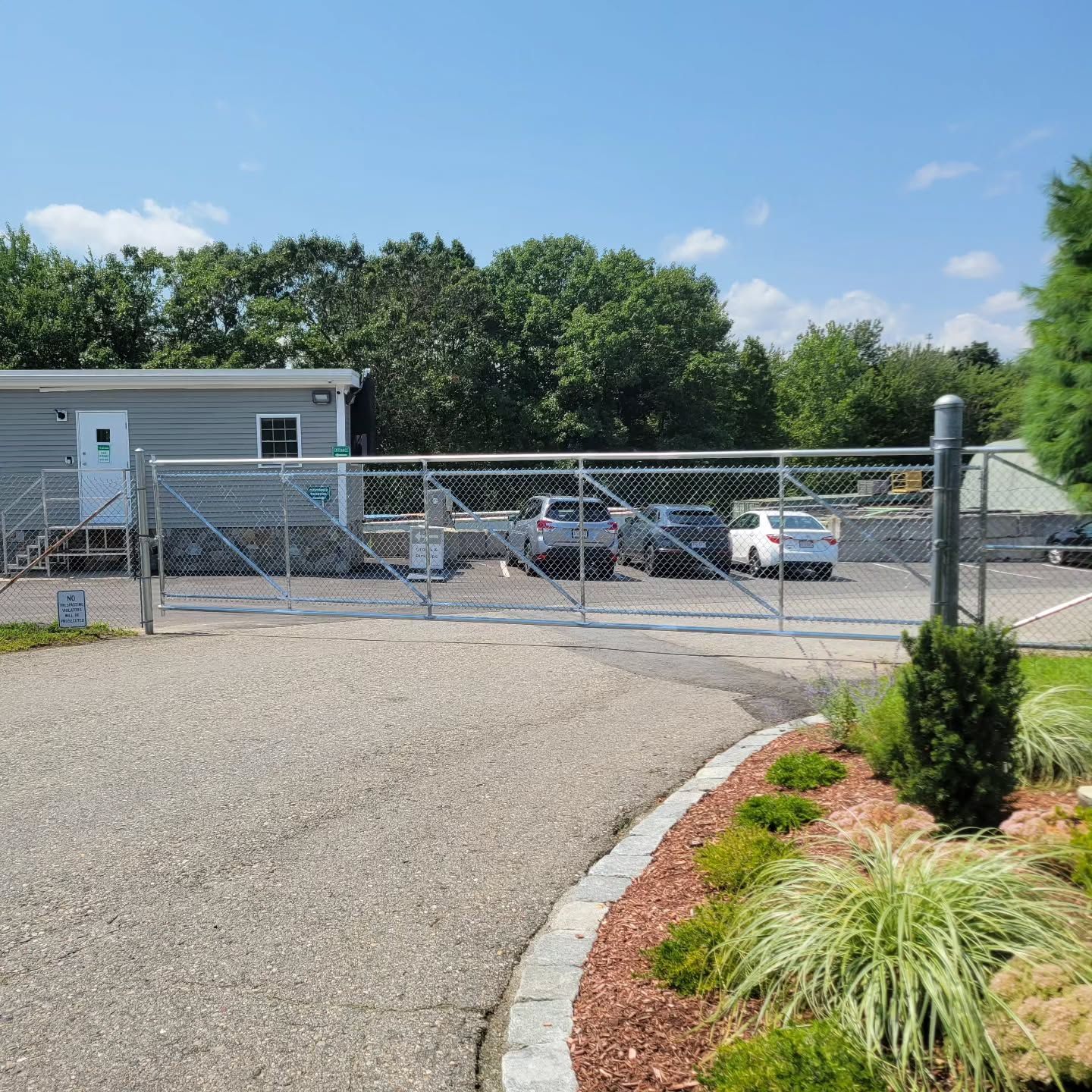 Chain-link fence gate entrance to a building. Cars are parked behind the gate; landscaping and blue sky visible.