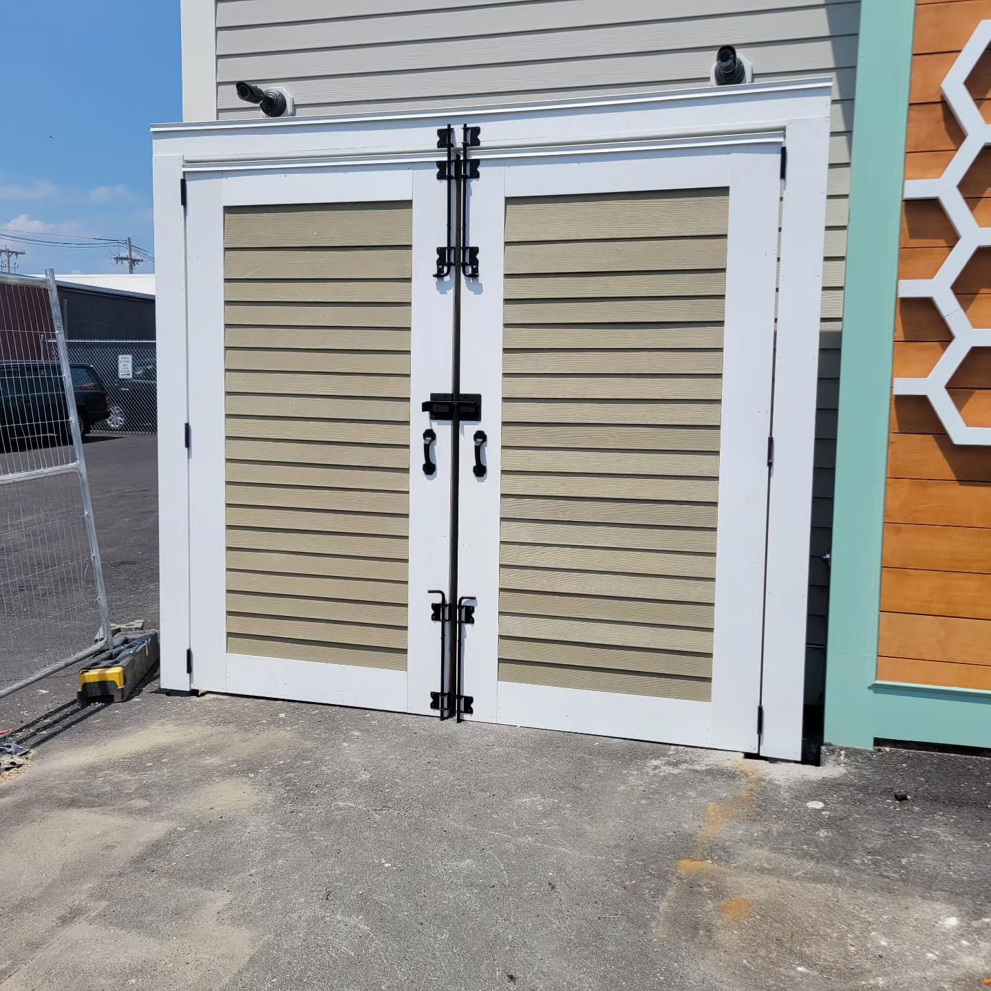 Double door with cream-colored trim and black hardware. Horizontal tan slats fill door panels. Set in concrete, with blue sky