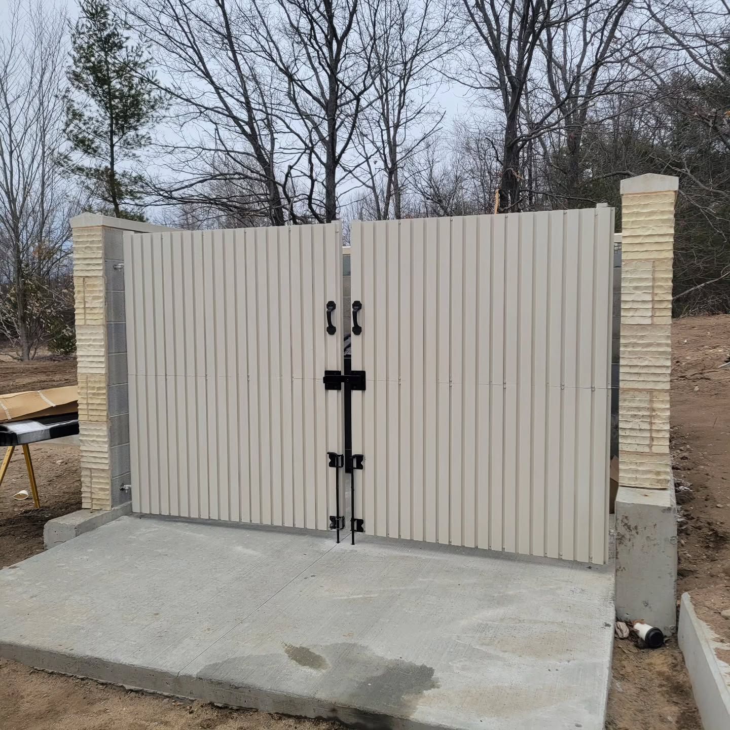 Beige vertical-slat gate with black hardware and stone pillars on a concrete pad.