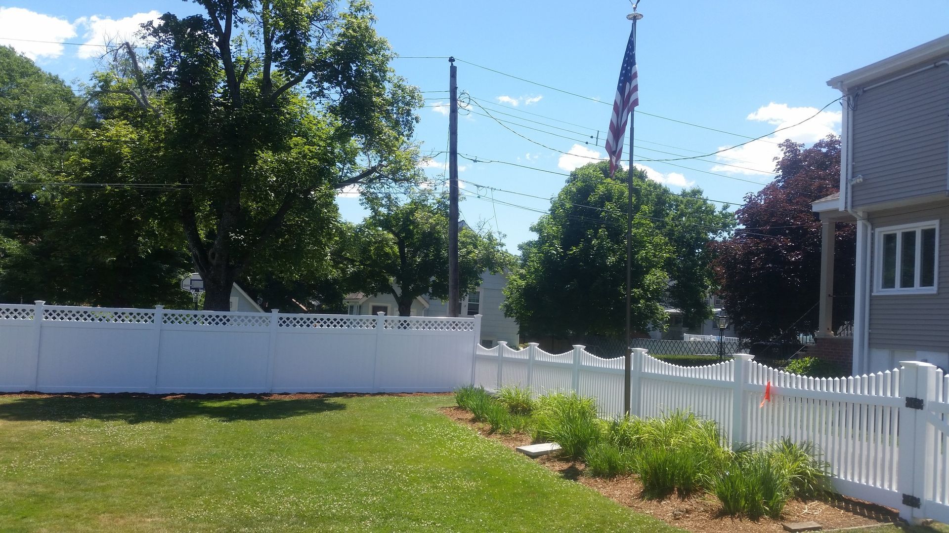 White picket fence along a green lawn with a tree and American flag under a bright blue sky.