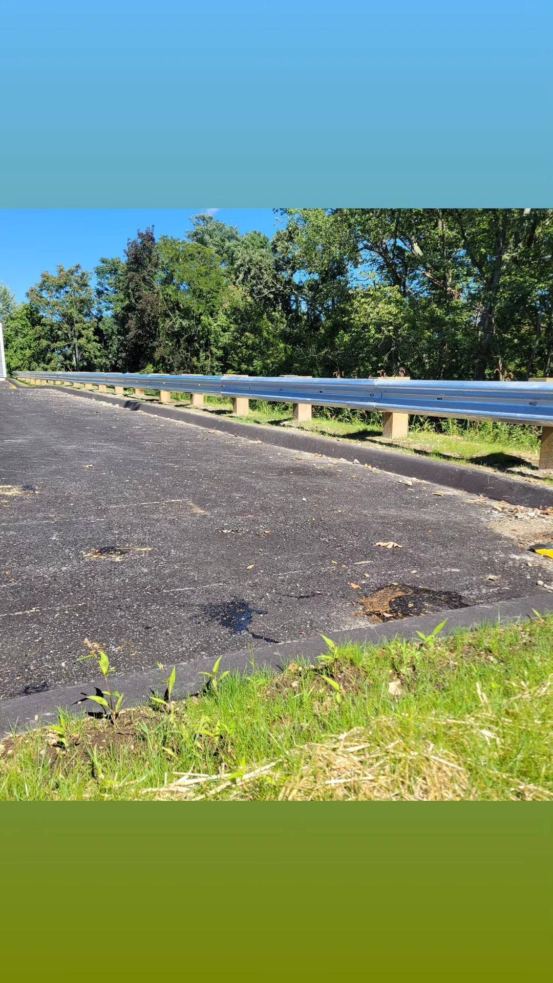 Bridge with a guardrail, asphalt road, and green trees against a bright blue sky.