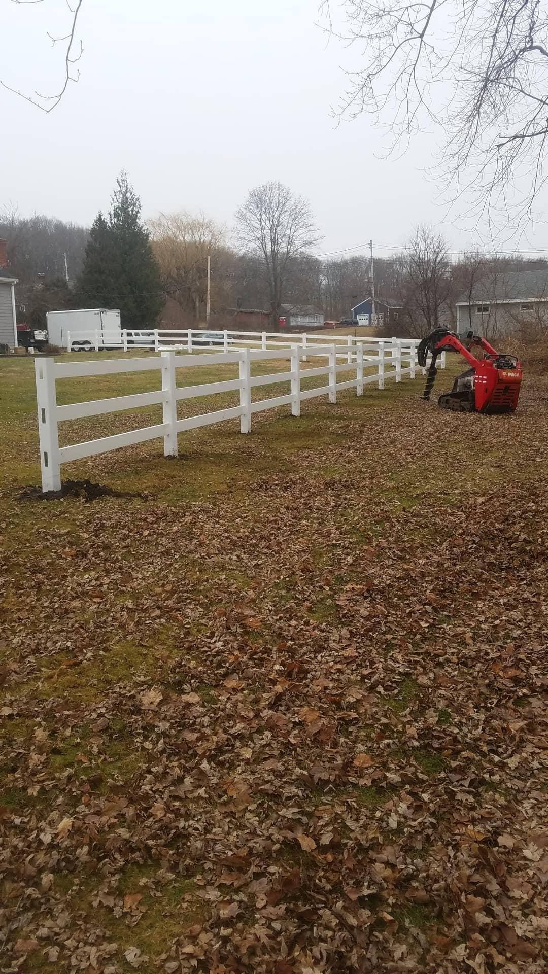 White fence in a field with a red tractor, trees, and scattered brown leaves.