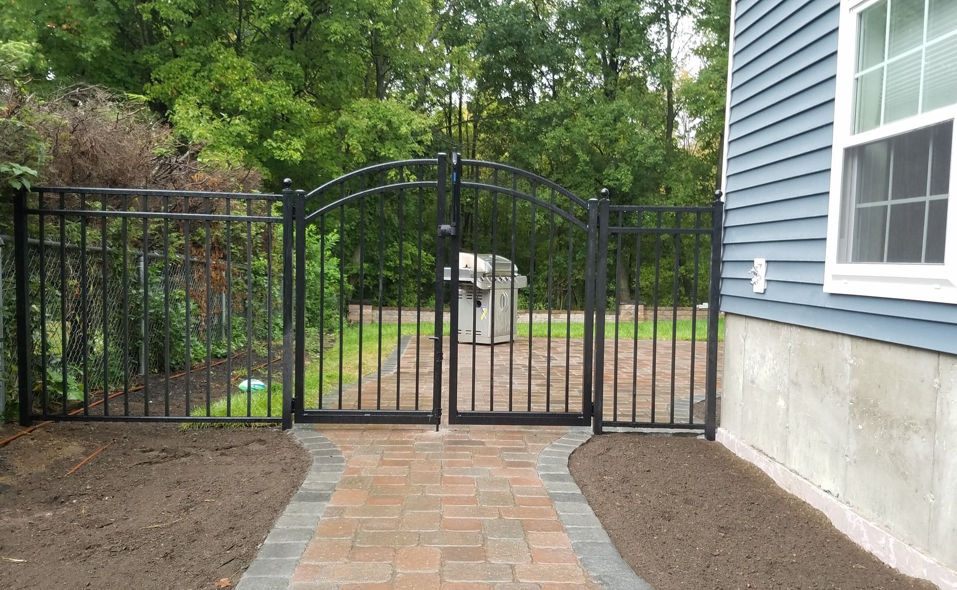 Black metal gate and fence along a brick pathway, next to a blue-sided house and green foliage.