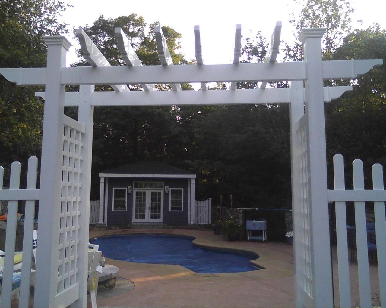 White pergola frames a view of a pool, small blue shed, and surrounding trees.