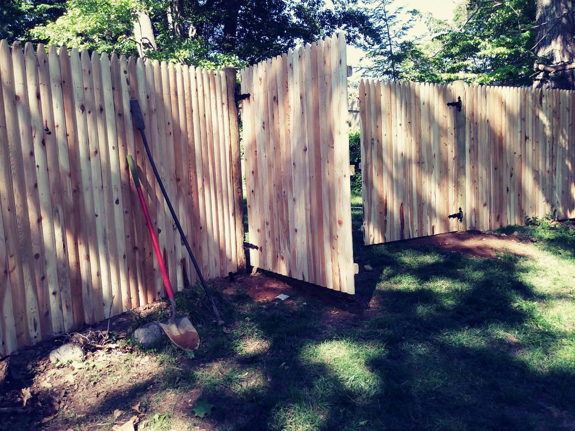 Wooden fence with open gate and tools leaning against the panels in a grassy yard.