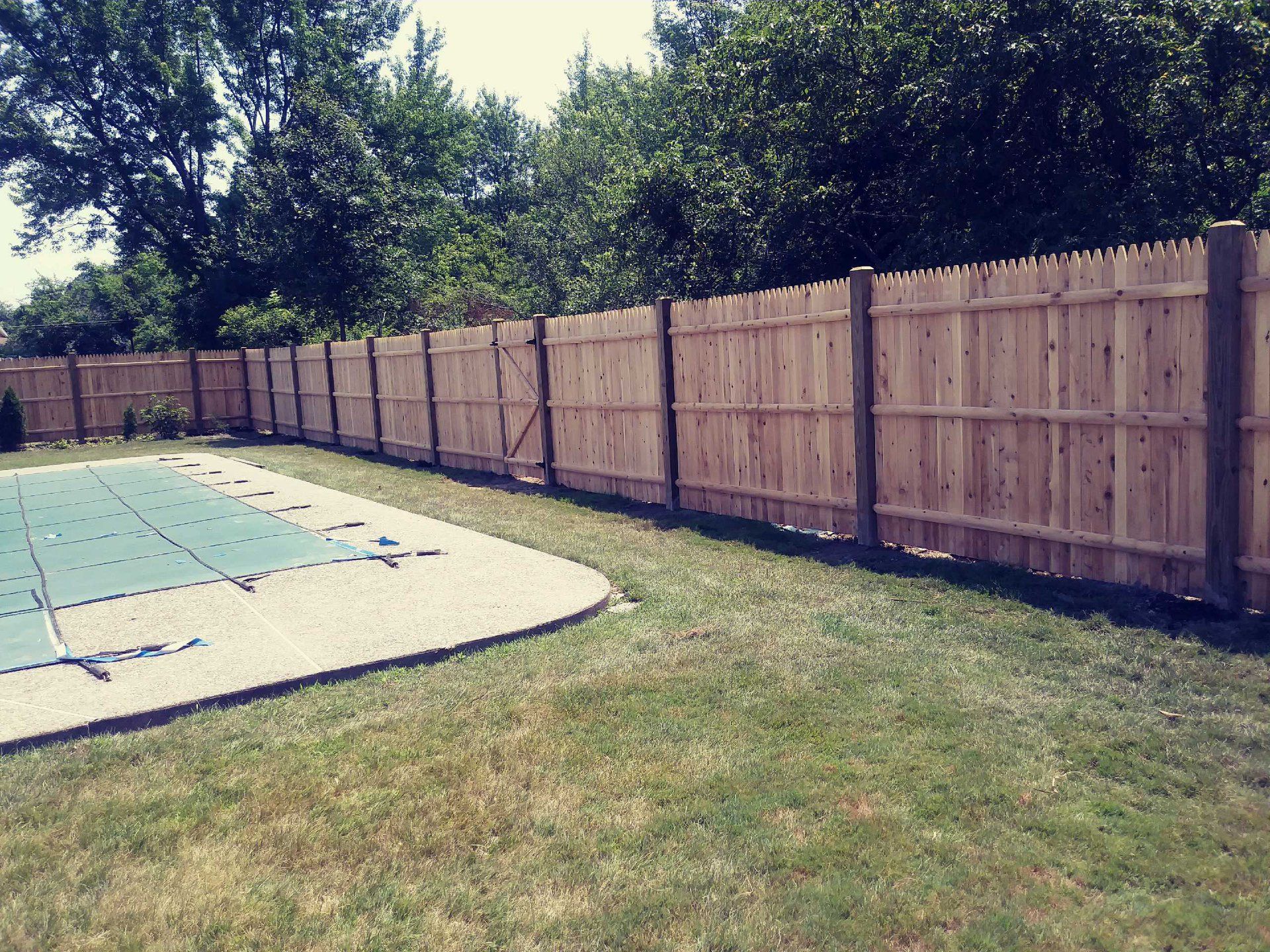 Wooden fence surrounds a backyard with a pool. Green grass and trees are in the background.