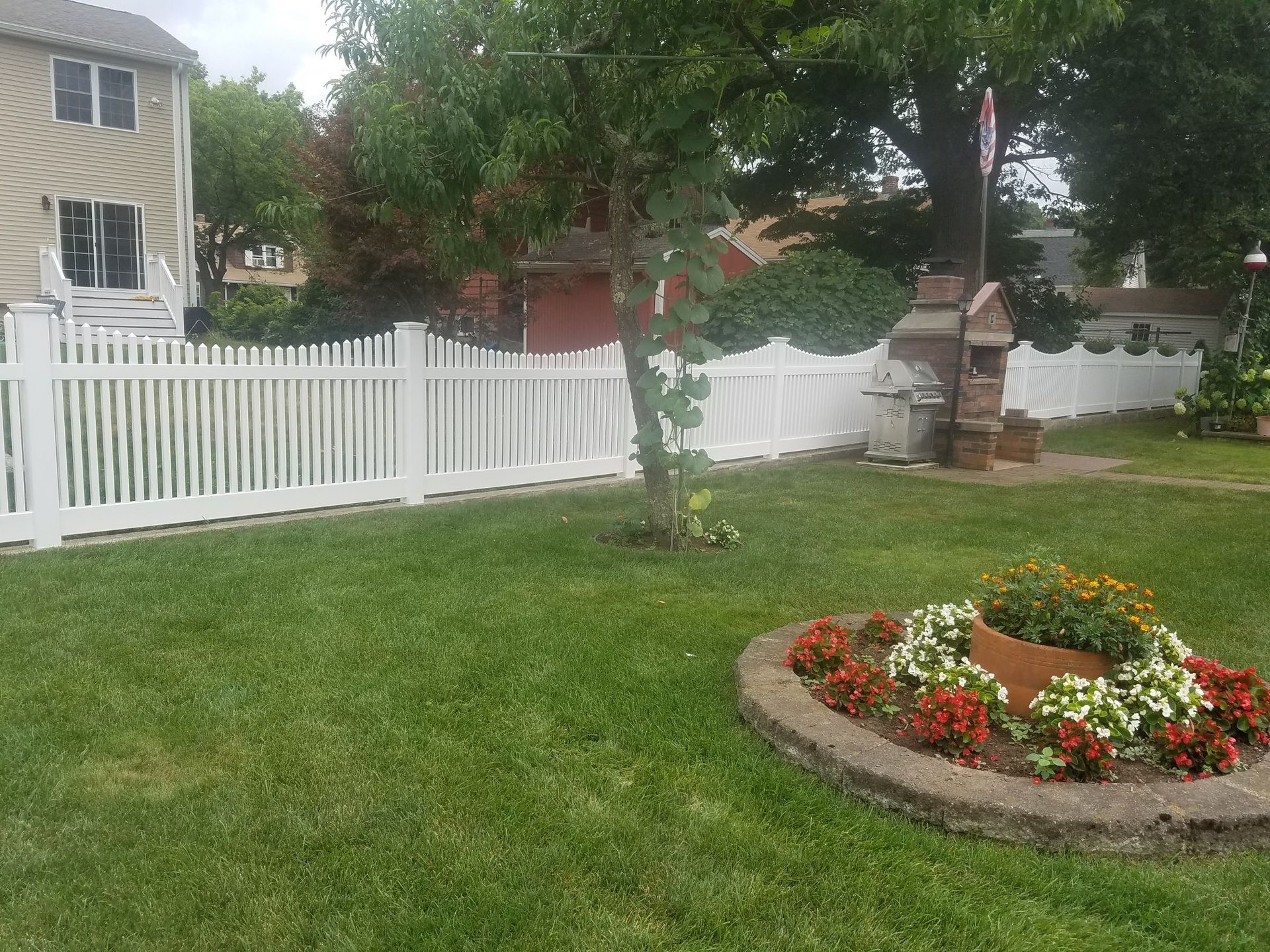 White picket fence encloses a green lawn with a flower bed and brick barbecue. A house is visible in the background.