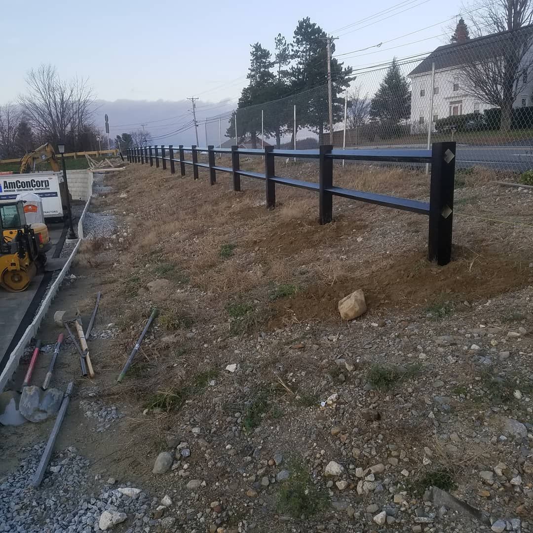 Black fence along a roadside, earth and construction equipment in foreground, building and trees in background.
