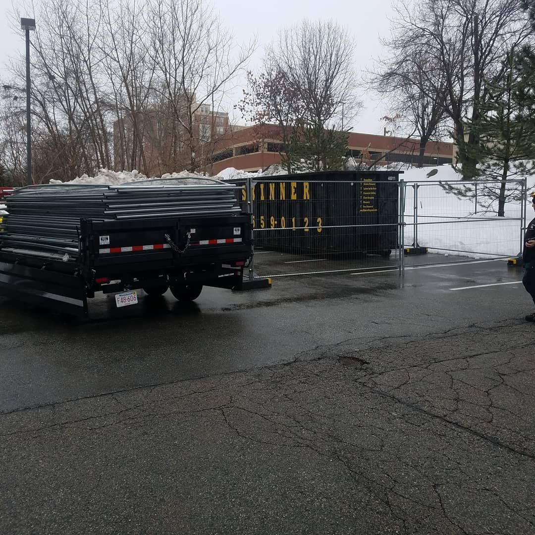 Black trailer loaded with metal and a dumpster blocked by a barrier on a wet asphalt lot near a building.