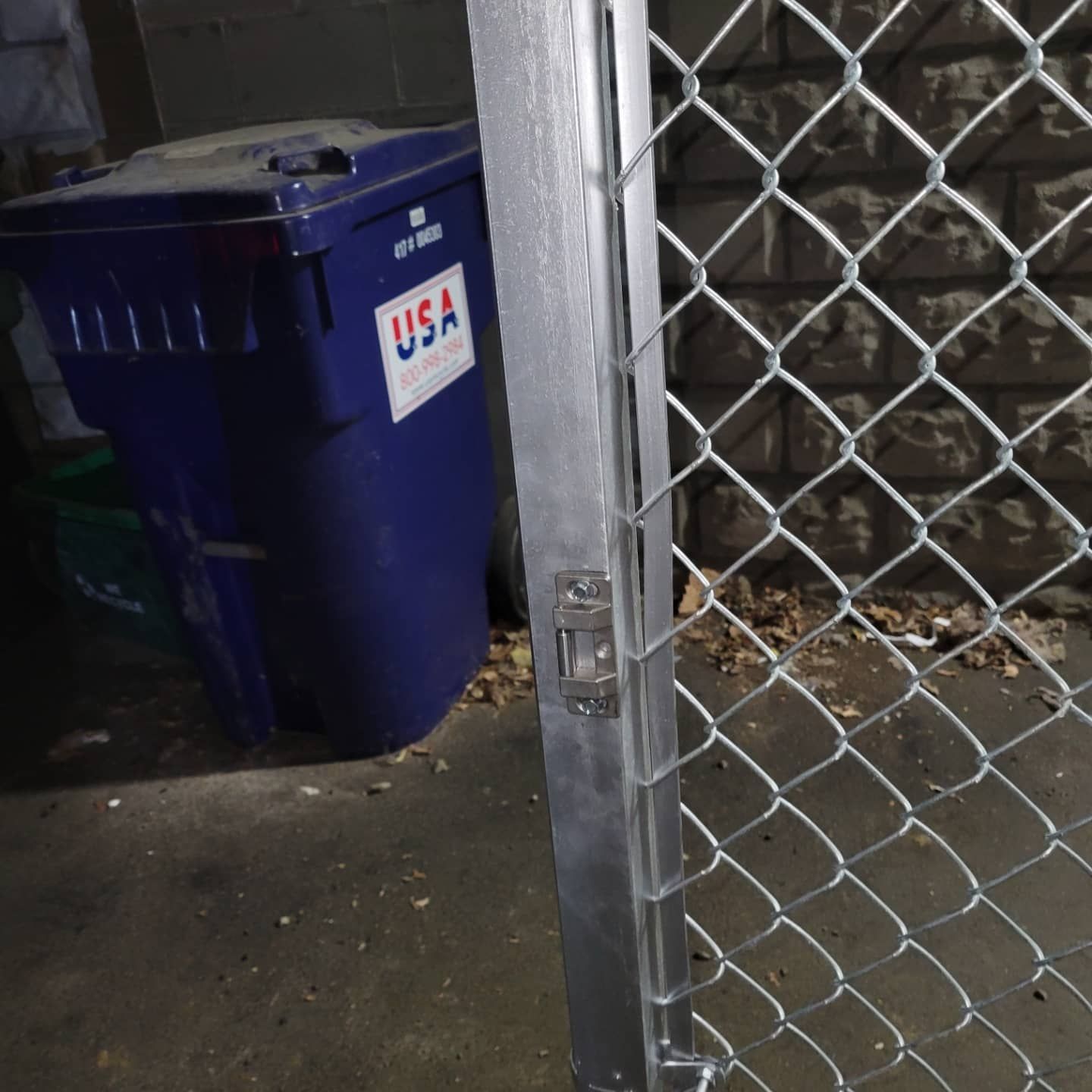 Blue trash bin beside a chain-link fence gate, in an area with a dirty floor.