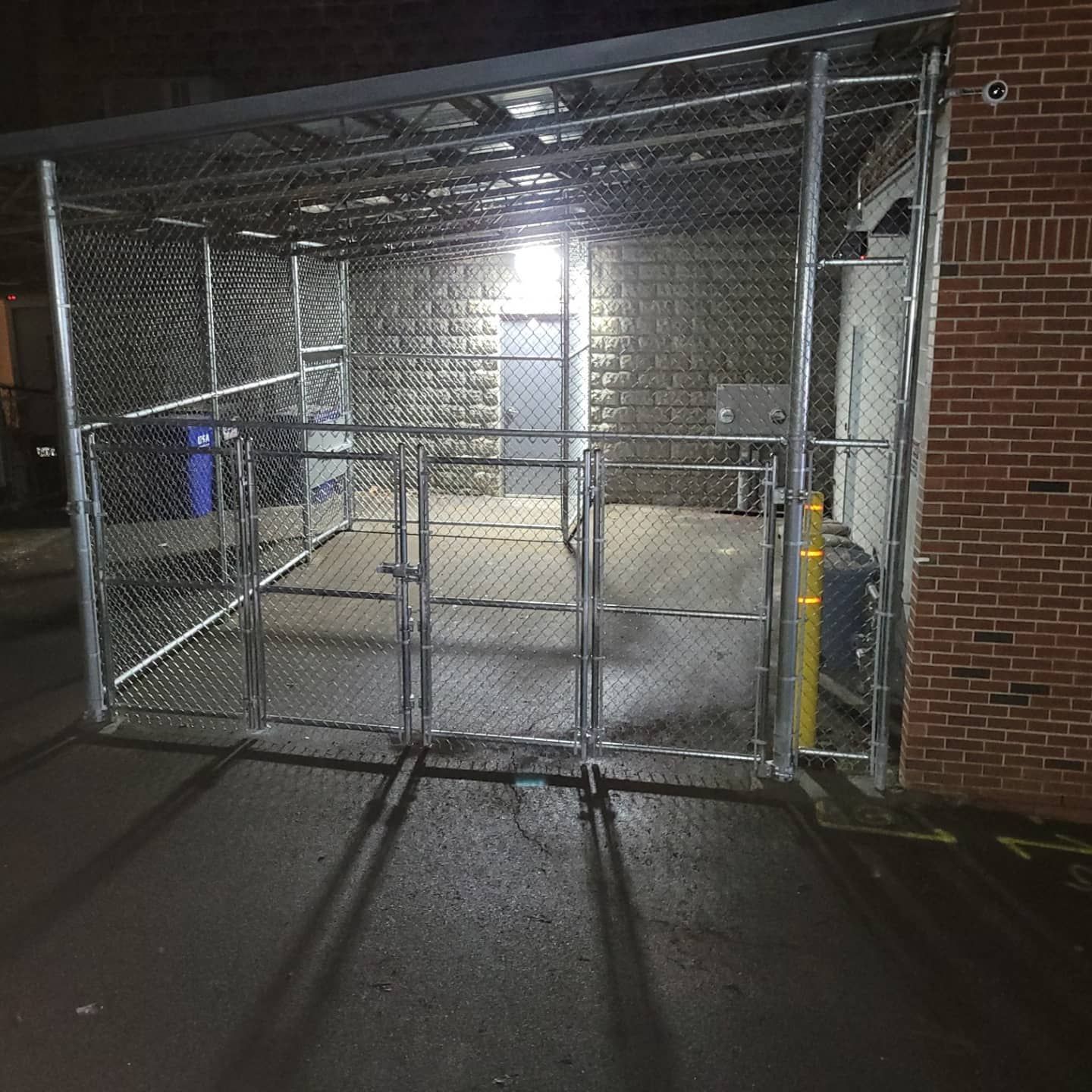 Chain-link fence enclosure with multiple gates, adjacent to a brick wall. Light from within illuminates the area.