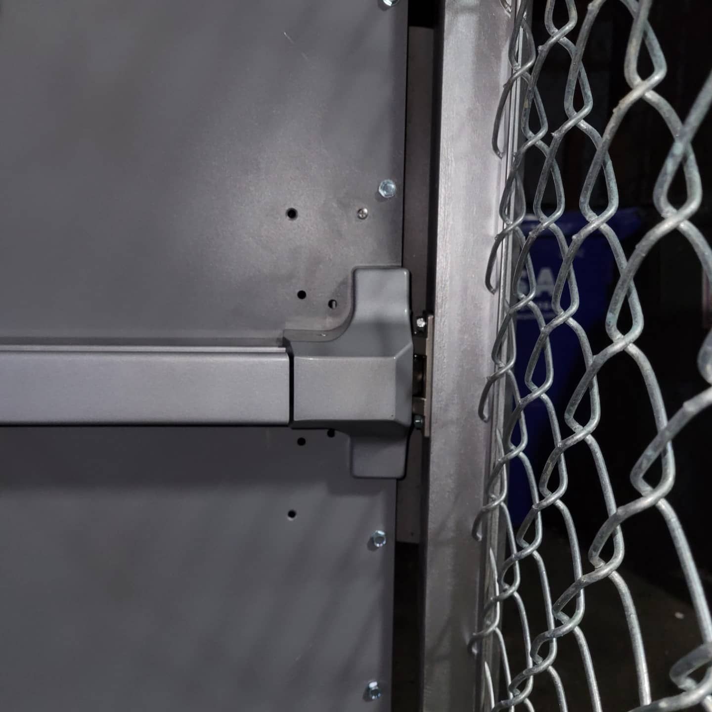 Gray metal door with push bar, chain-link fence on the right.