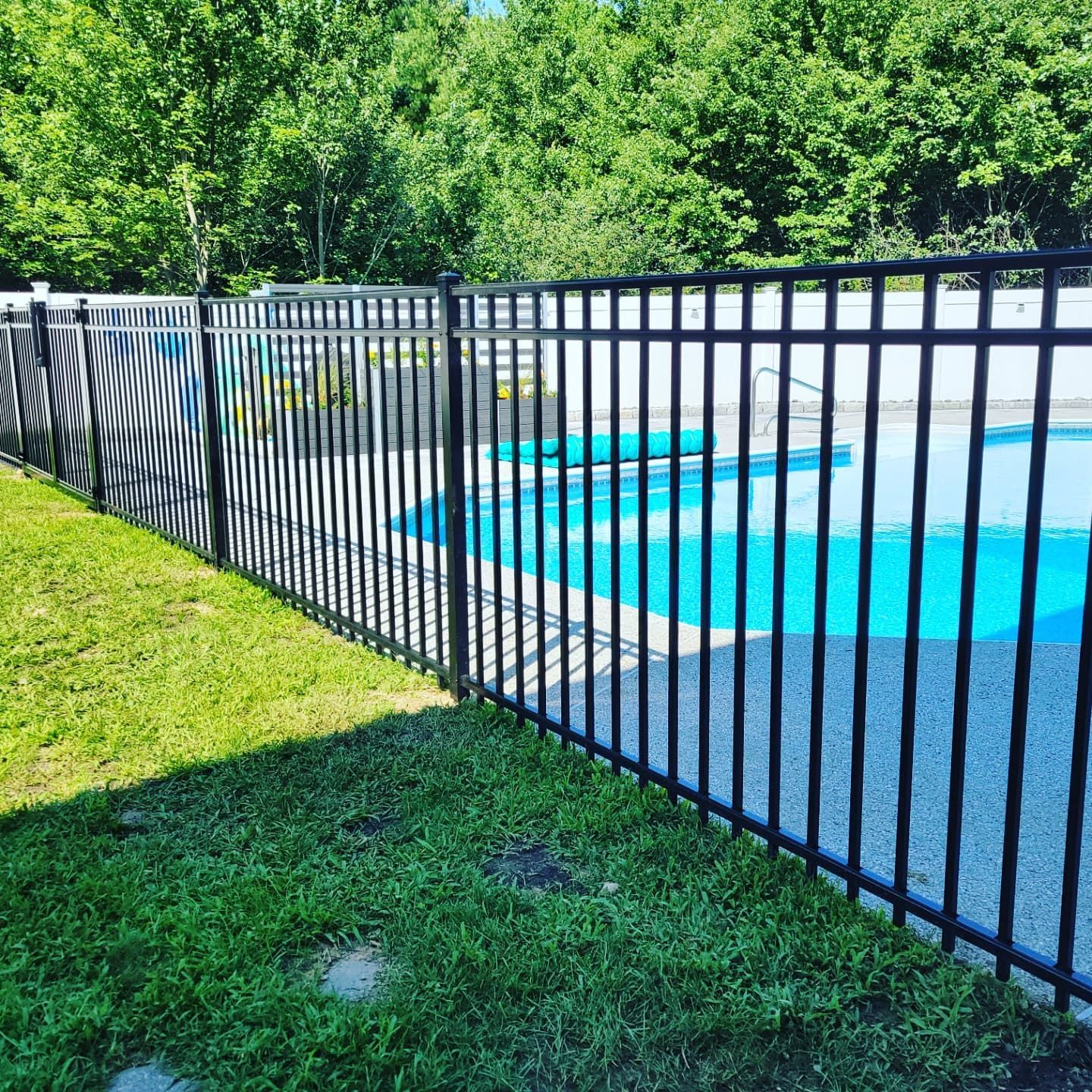 Black metal fence around a blue swimming pool in a grassy yard, trees in the background.