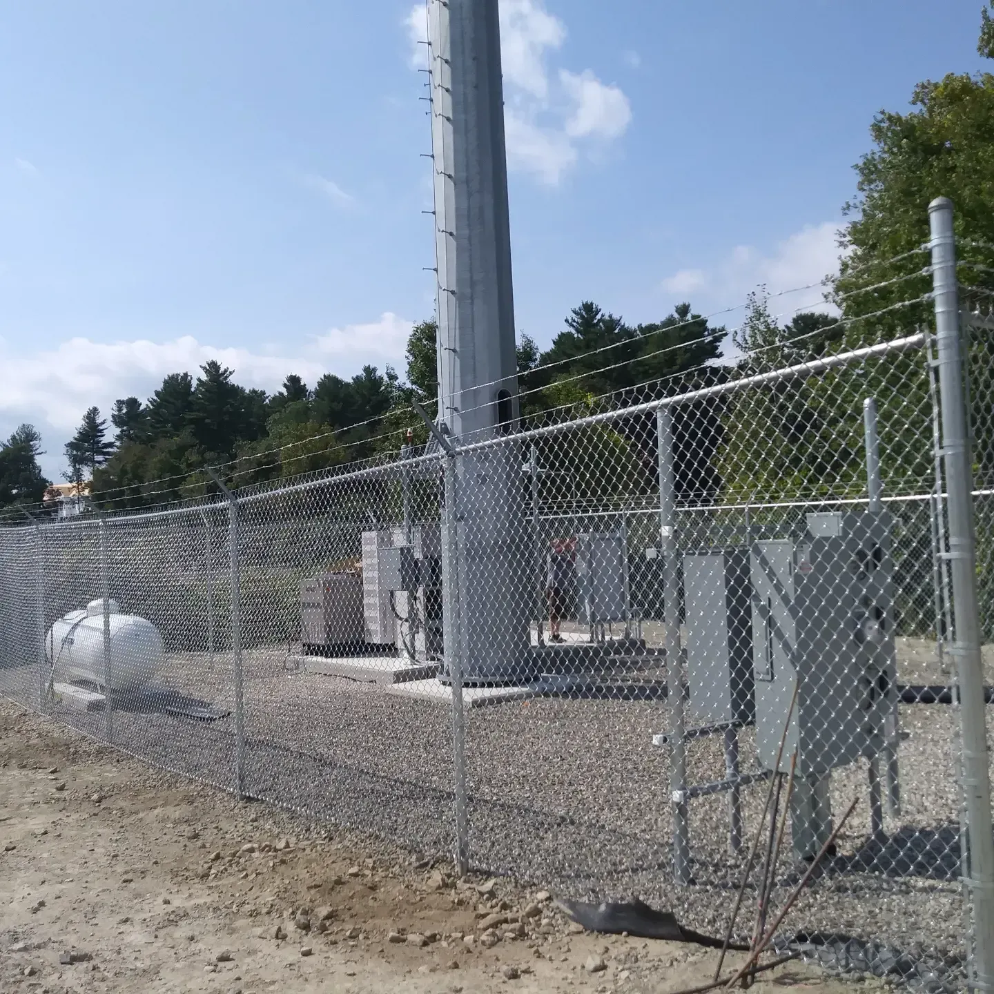 A fenced-in cell tower on a gravel lot. A propane tank and gray utility boxes are visible.