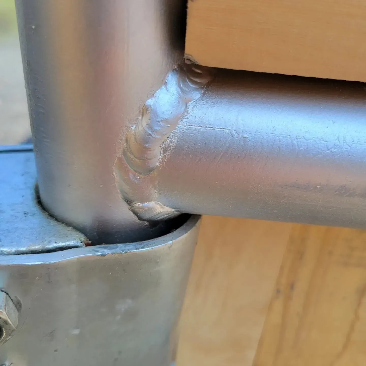 Close-up of a silver metal tube welded to another tube, with a wooden surface behind it.