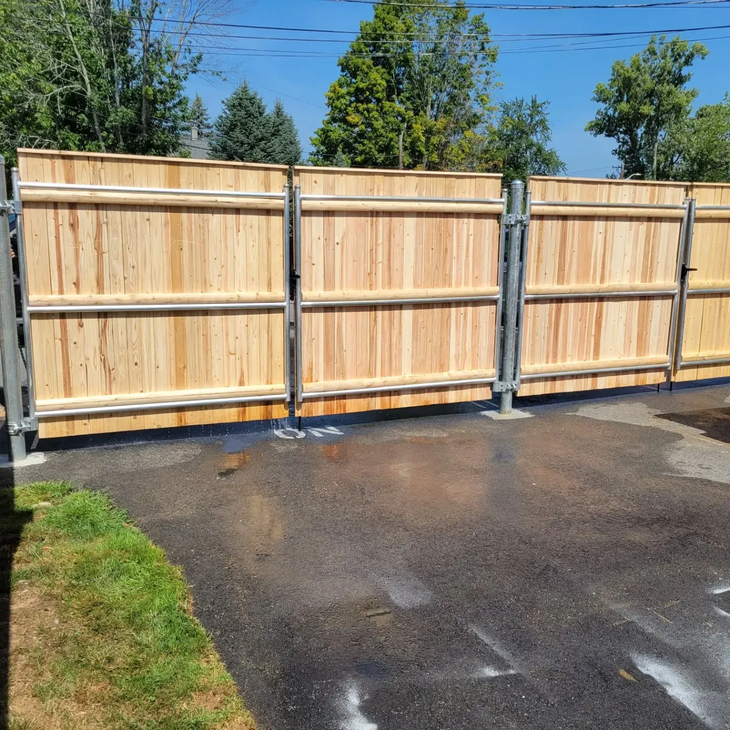 Wooden fence with metal posts on asphalt driveway, with grass and trees in the background.