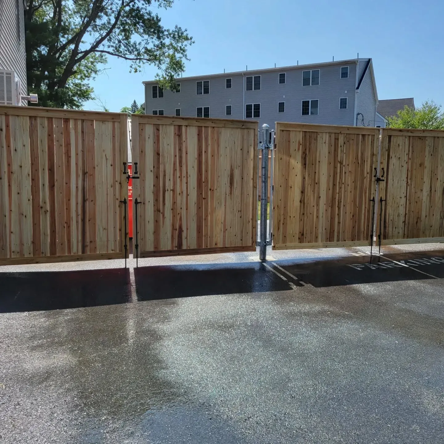 Wooden fence with a gate, on a paved surface. Buildings and a tree are in the background.