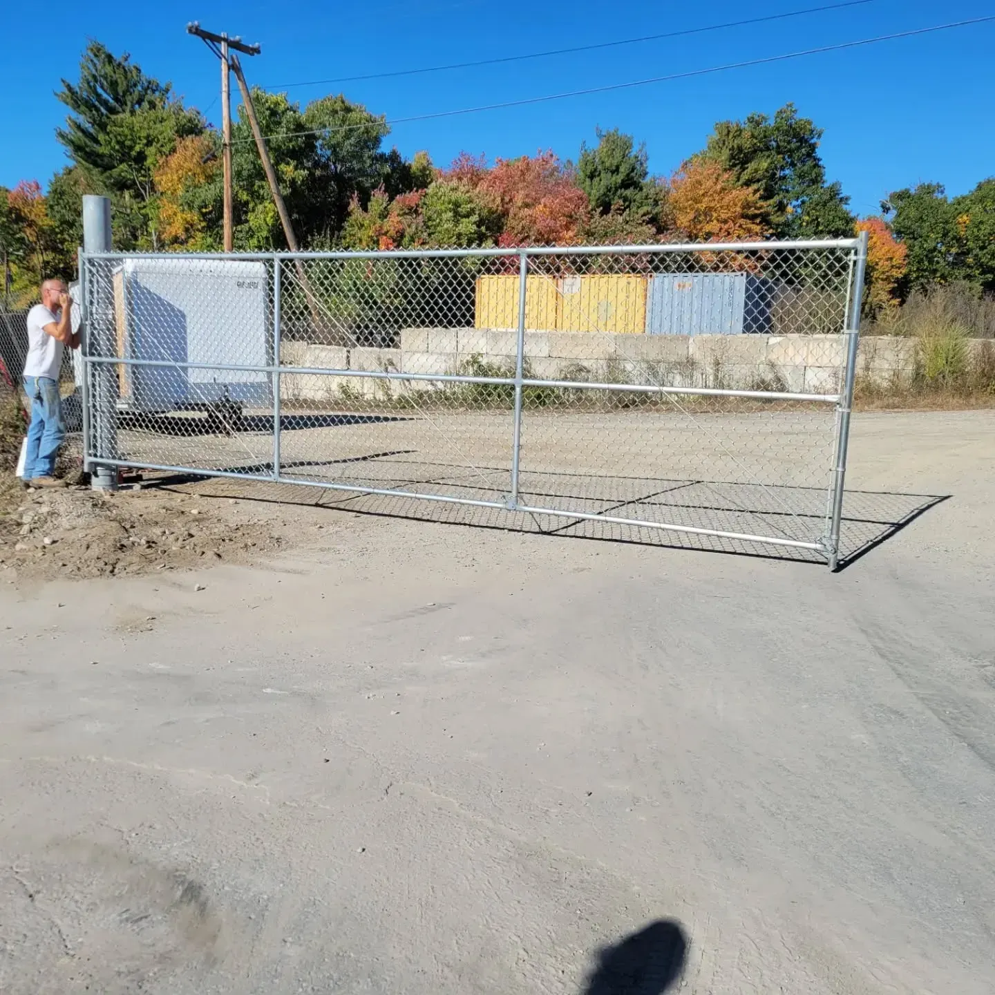 Man adjusting a chain-link fence gate on a paved surface. Fall foliage in background.