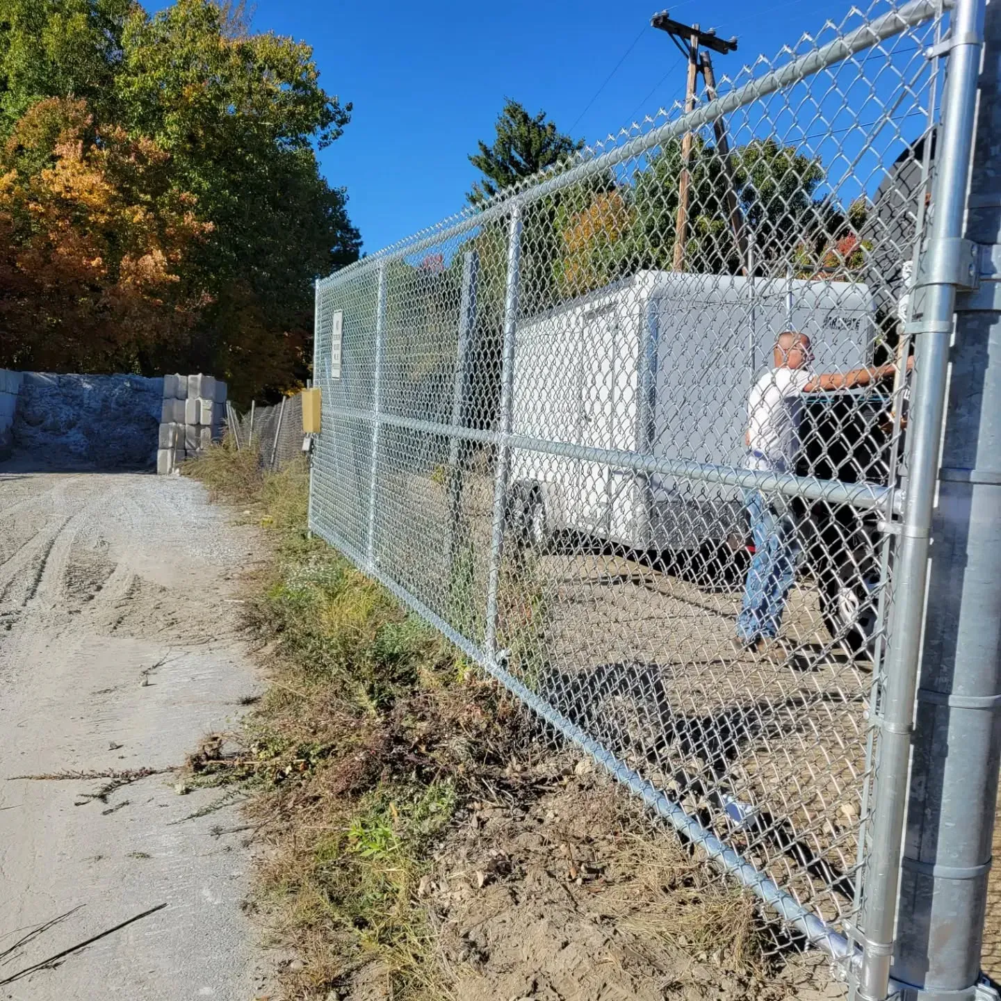 Chain-link fence in front of a gravel road, person holding the fence. White storage container in the background, blue sky.