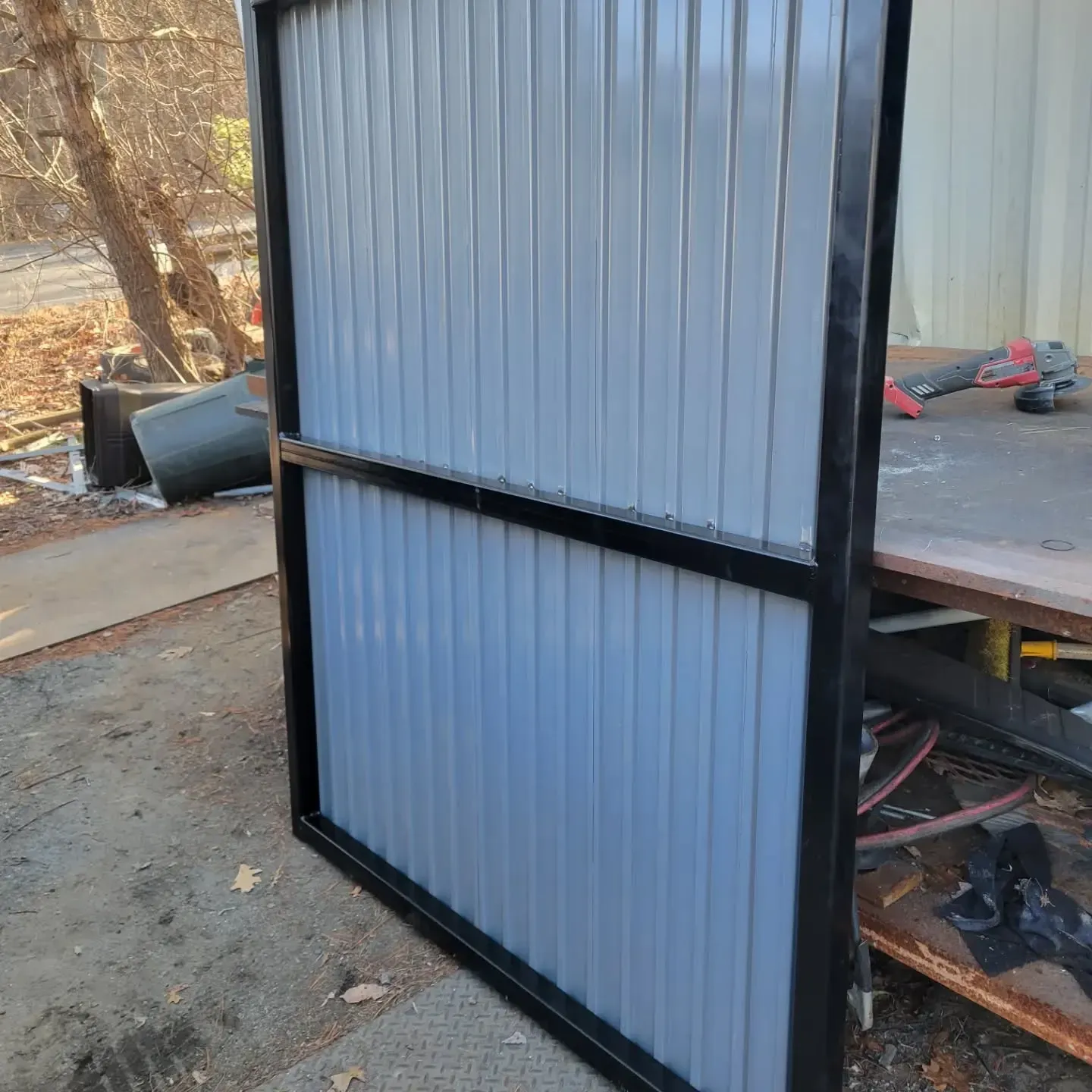 Black metal frame with corrugated gray metal panels. Outdoors, next to a workbench.