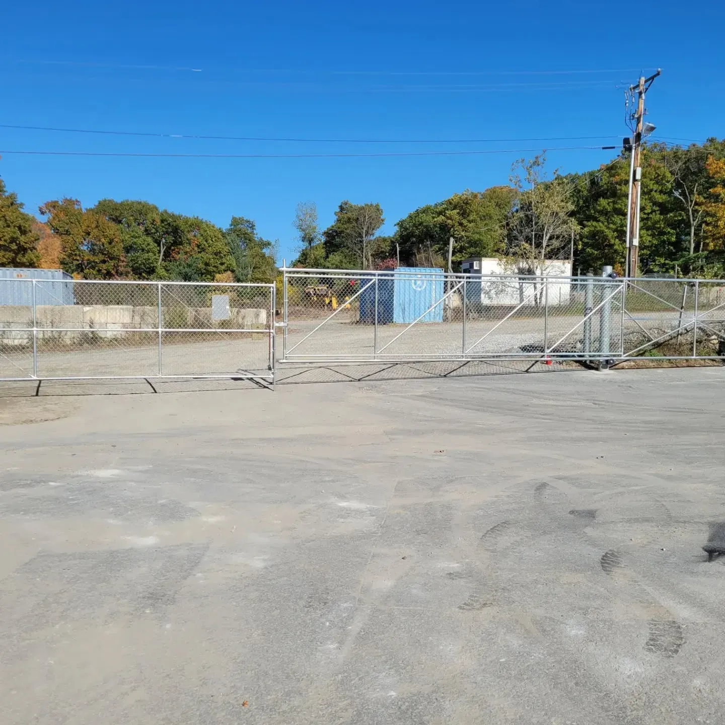 Chain-link fence with an open gate on a gray paved surface, trees, and blue sky.