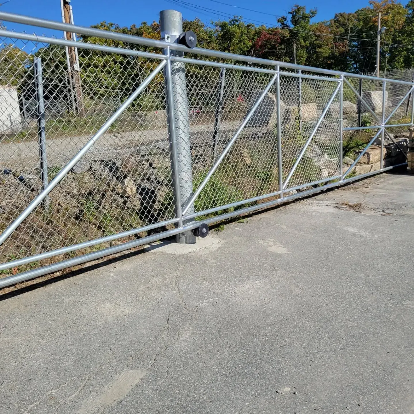 A metal chain-link gate. The gate is closed and has diagonal supports, set on concrete, in front of foliage.