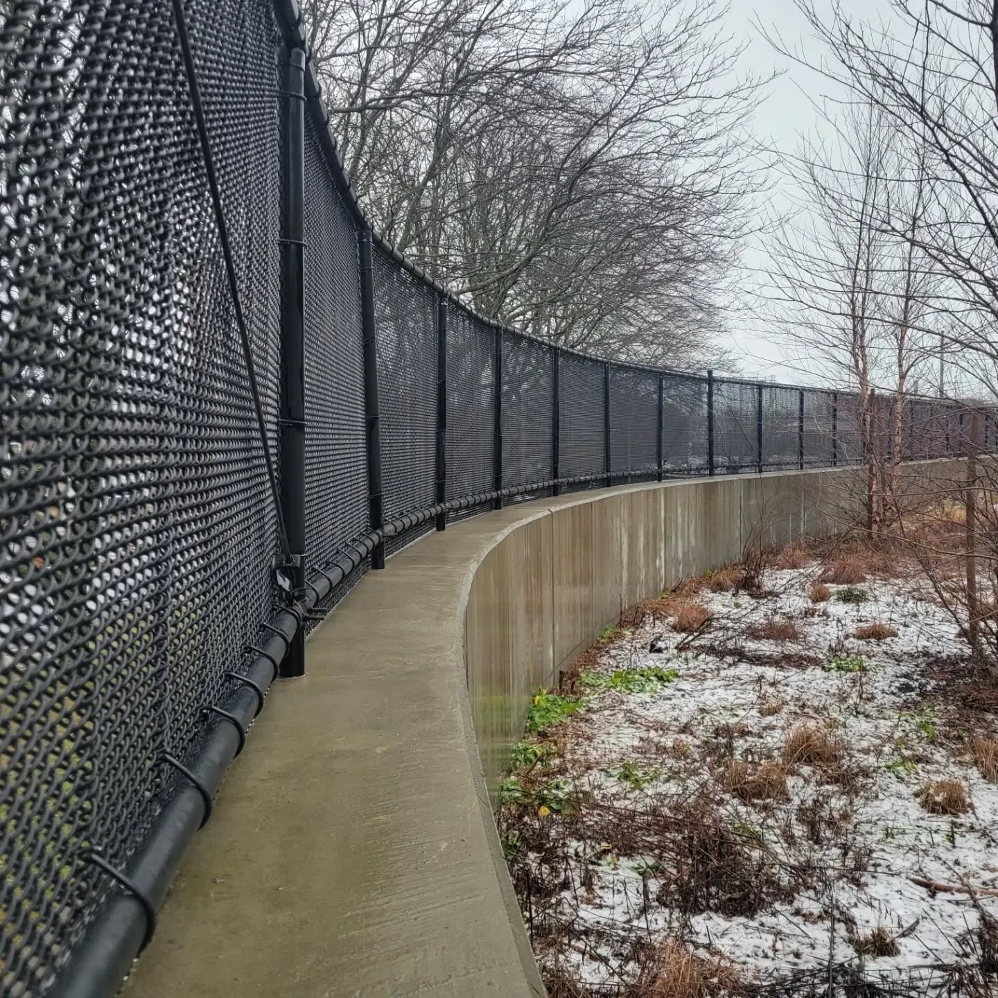 Curving black fence atop a concrete barrier, along a path with sparse vegetation. Overcast sky.