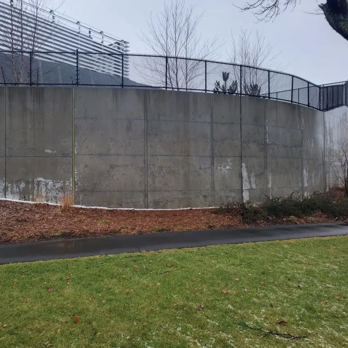 Concrete wall with a chain-link fence on top. Path and grass in foreground. Cloudy day.